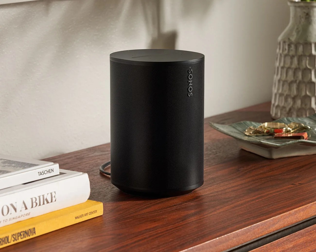 Black cylindrical Sonos speaker on a wooden surface next to stacked books and a decorative dish.