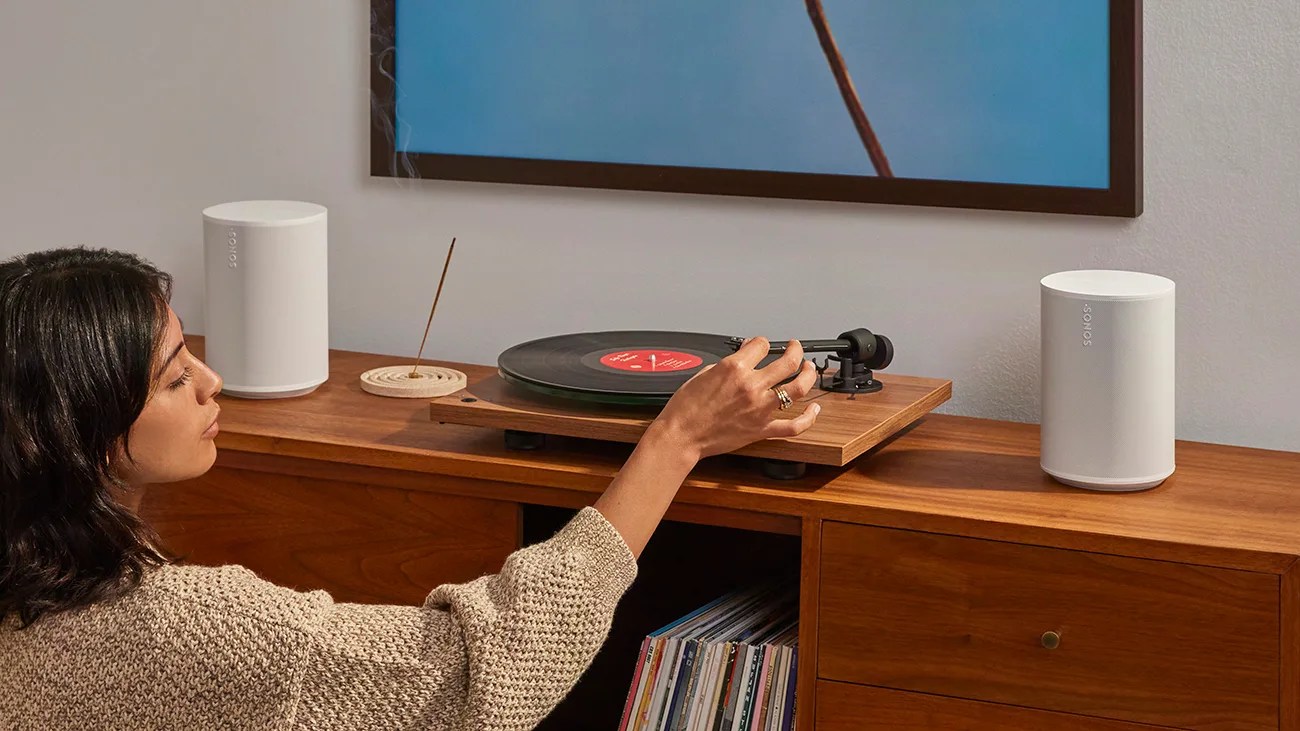 Woman adjusting tonearm on wooden turntable with two white Sonos speakers on a wooden cabinet.