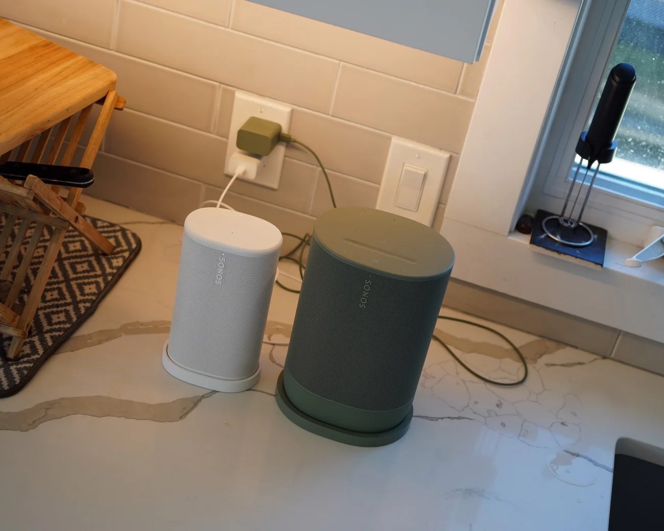 Two Sonos speakers, one white and one green, placed on a kitchen countertop near a power outlet.