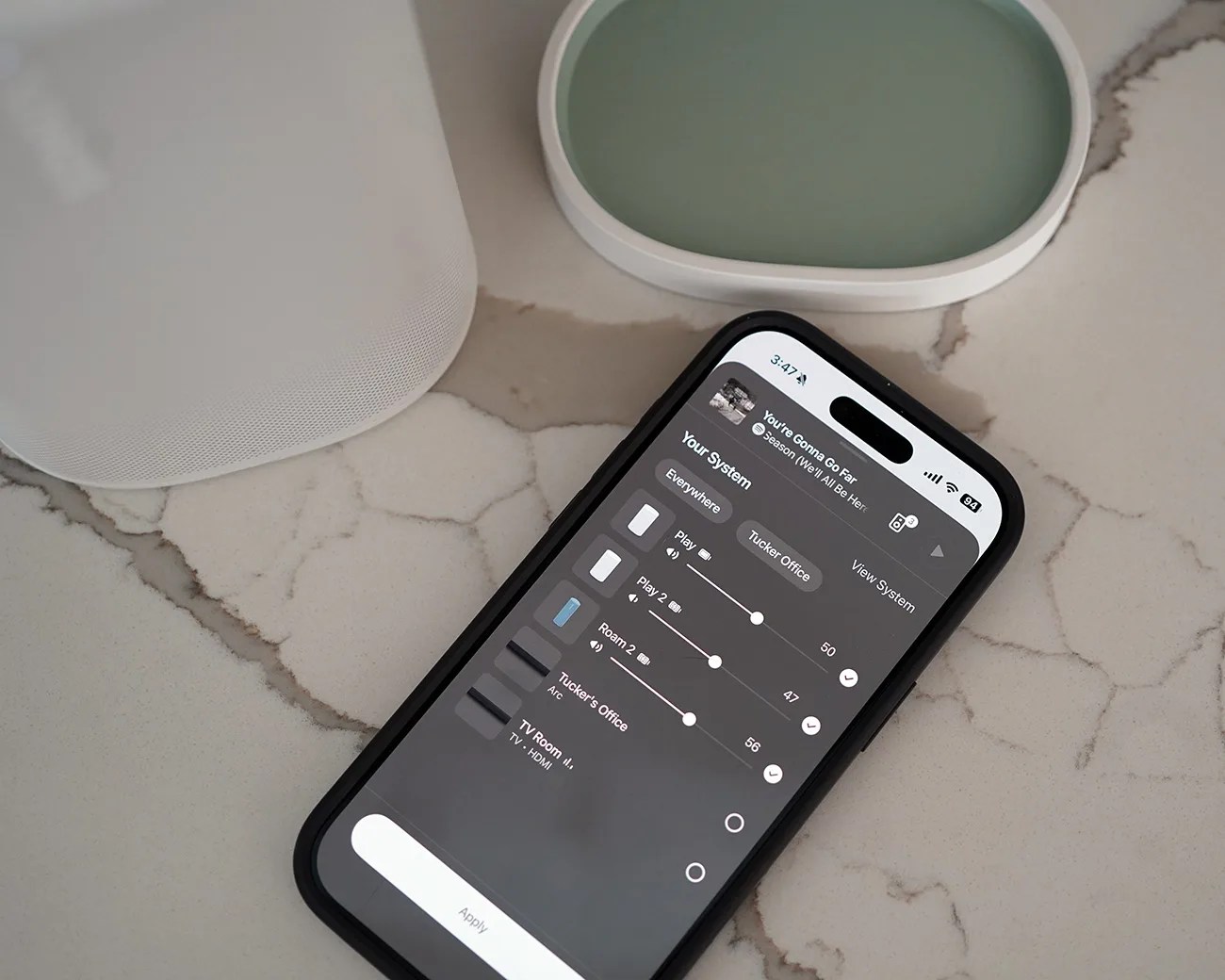 Smartphone with a black case displaying a home audio system control app on a marble countertop next to a white speaker and a wireless charger.