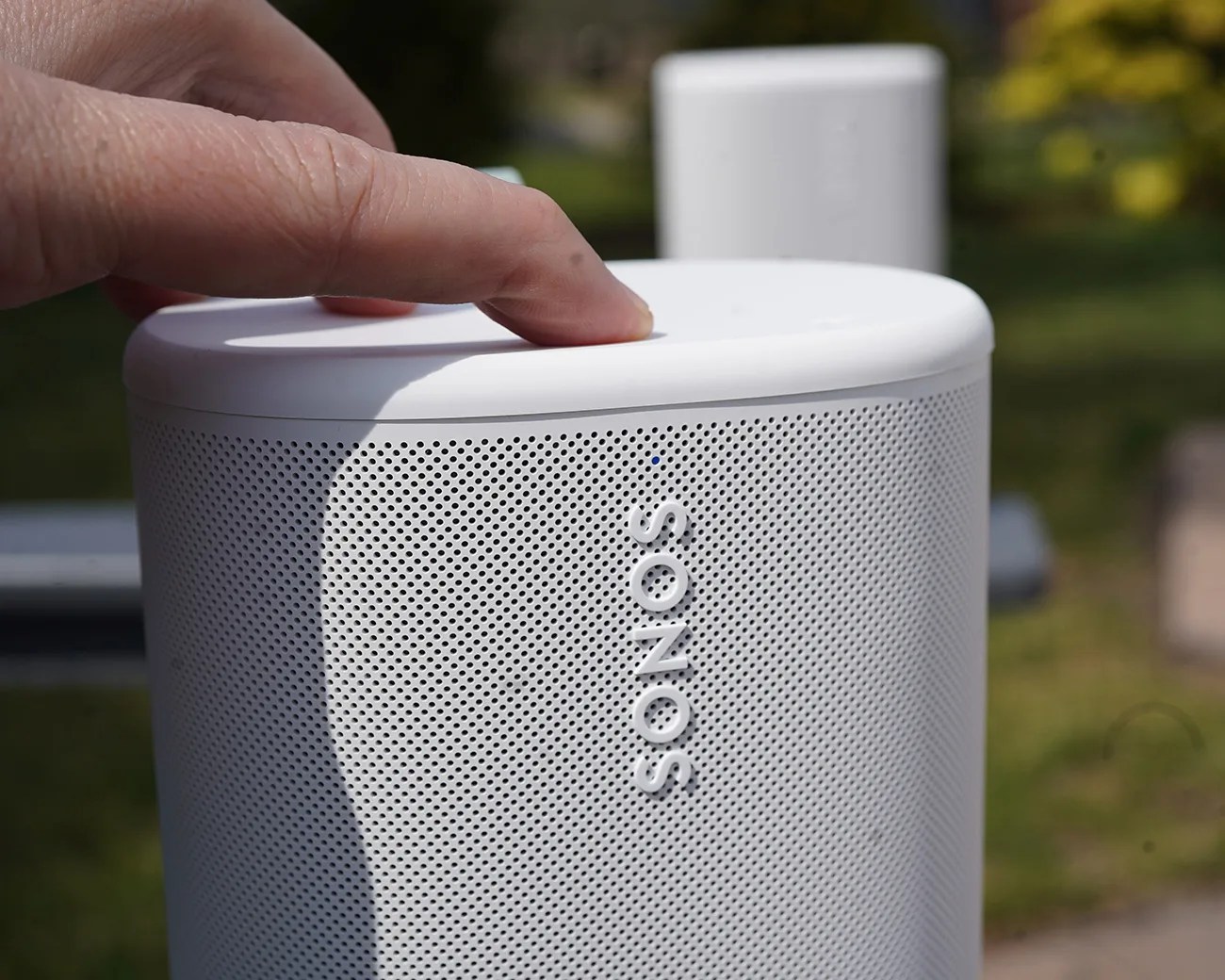 White Sonos speaker with perforated grille and a finger pressing a button on top.