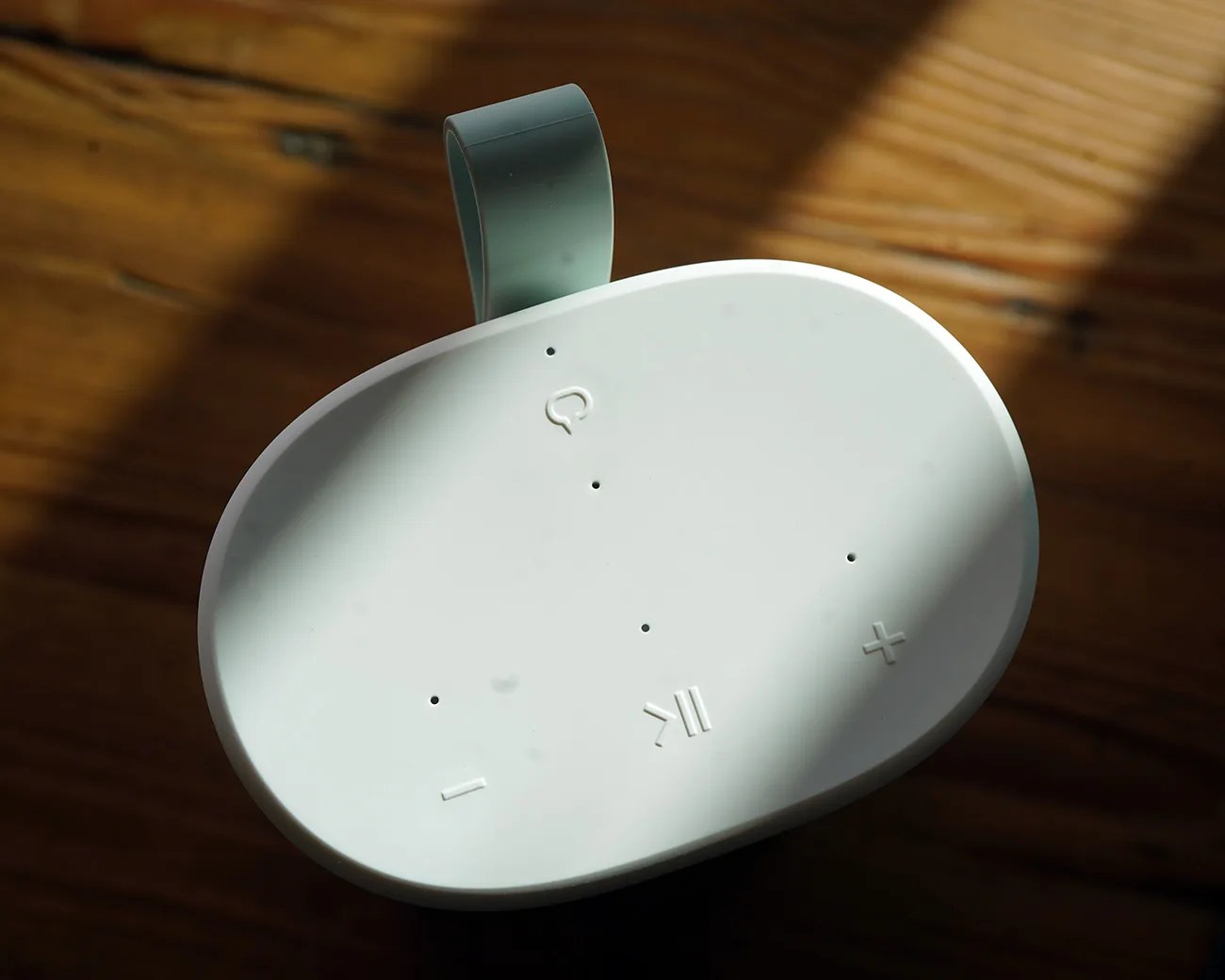 White oval Bluetooth speaker with control buttons and a gray loop handle on a wooden surface.