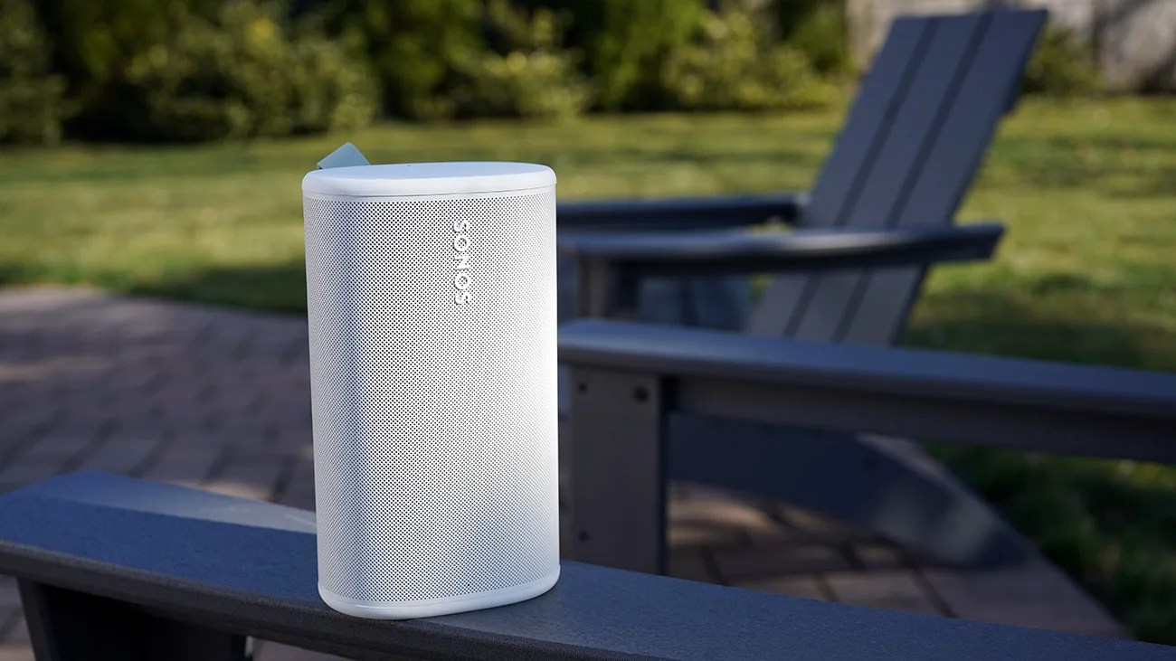 White Sonos wireless speaker placed on the armrest of a dark outdoor chair with grass in the background.