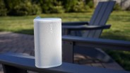 White Sonos wireless speaker placed on the armrest of a dark outdoor chair with grass in the background.