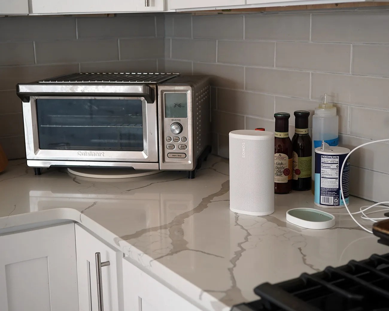 Stainless steel Cuisinart toaster oven on a marble countertop next to a white Sonos speaker and condiment bottles.