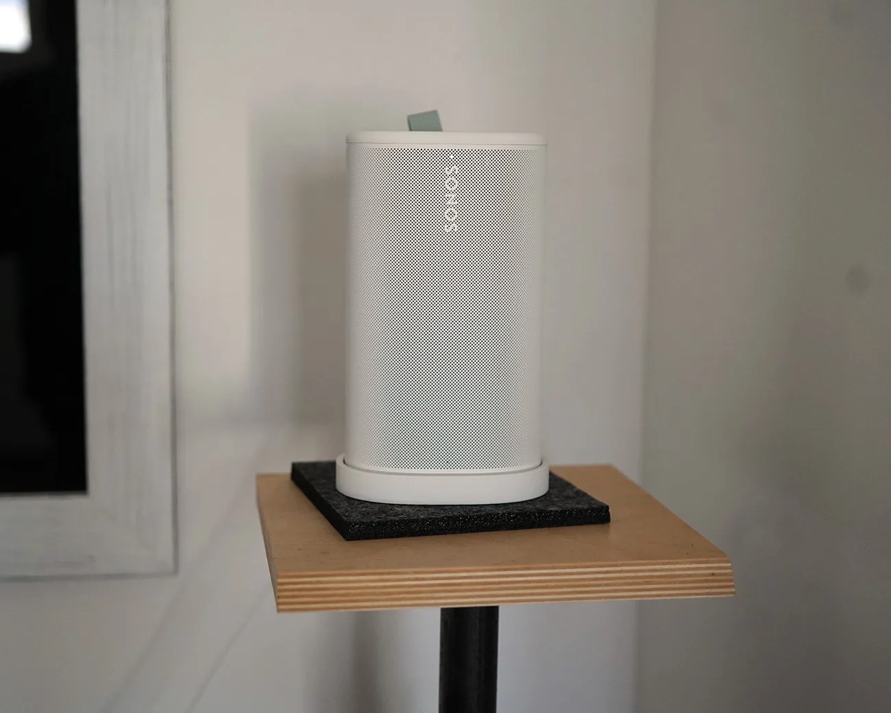White Sonos speaker on a black felt pad atop a wooden stand against a light wall.