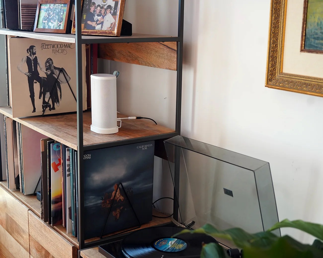 White cylindrical wireless speaker on a wooden shelf with vinyl records and a record player nearby.
