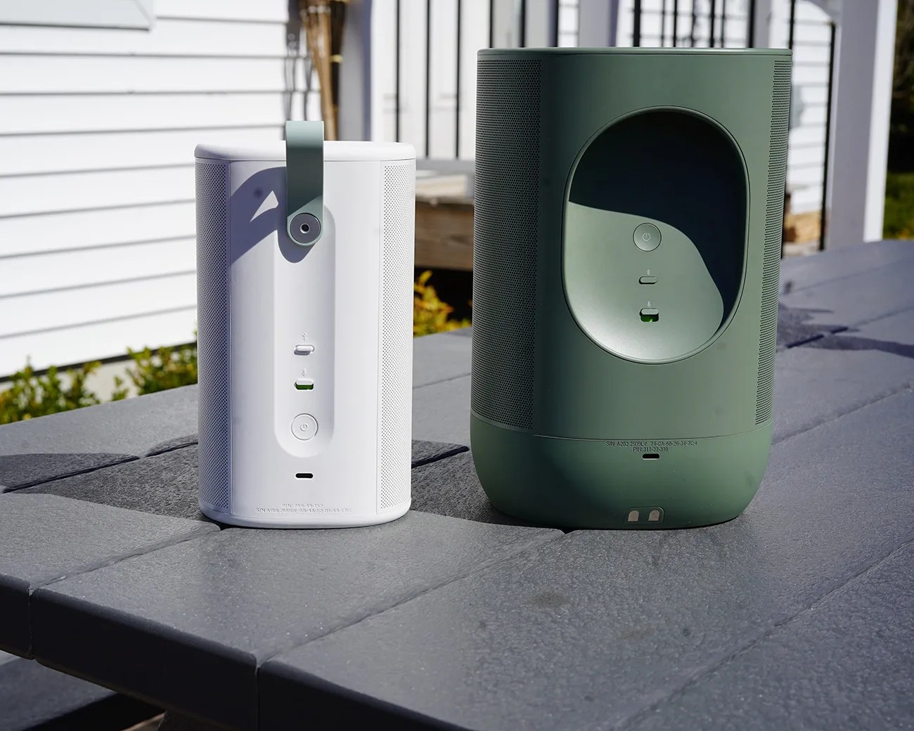 Two portable speakers, one white with a green handle and one larger green, placed on a gray outdoor table.