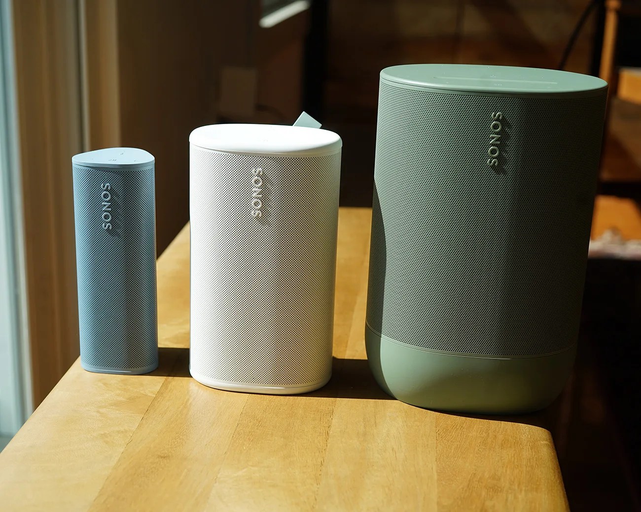 Three Sonos wireless speakers in different sizes and colors (blue, white, and green) on a wooden table.
