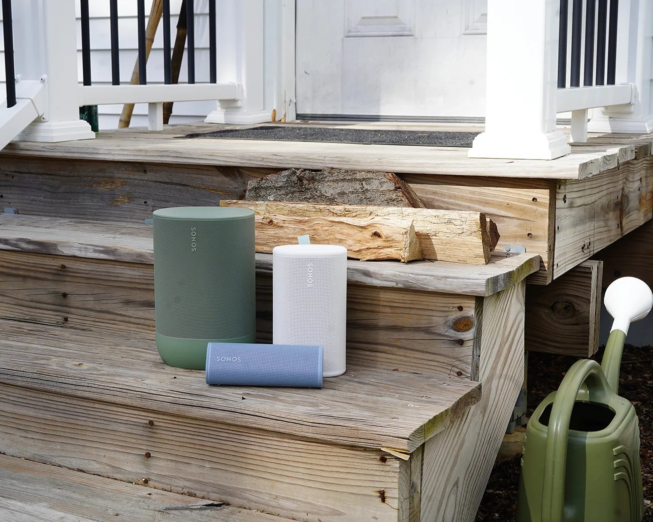 Three Sonos speakers in green, white, and blue placed on wooden porch steps next to a green watering can.