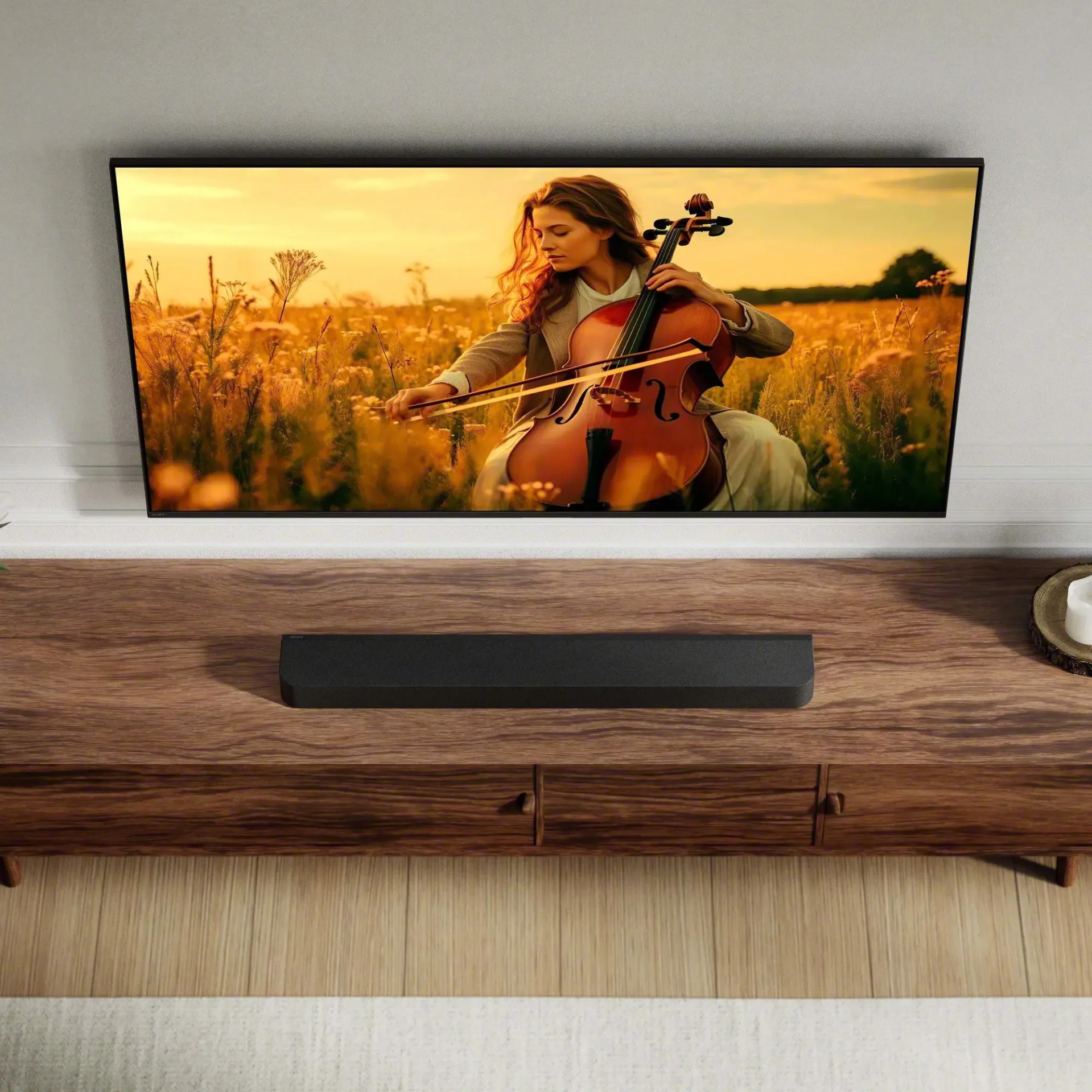 Black rectangular soundbar on a wooden TV stand below a wall-mounted flat-screen TV showing a woman playing cello in a field.