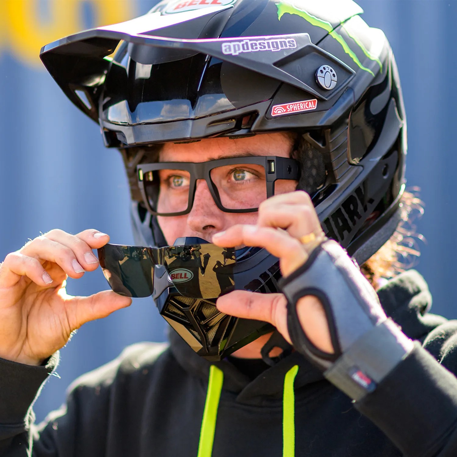 Person wearing a black Bell helmet with green accents and black glasses holding a tinted visor in front of their face.
