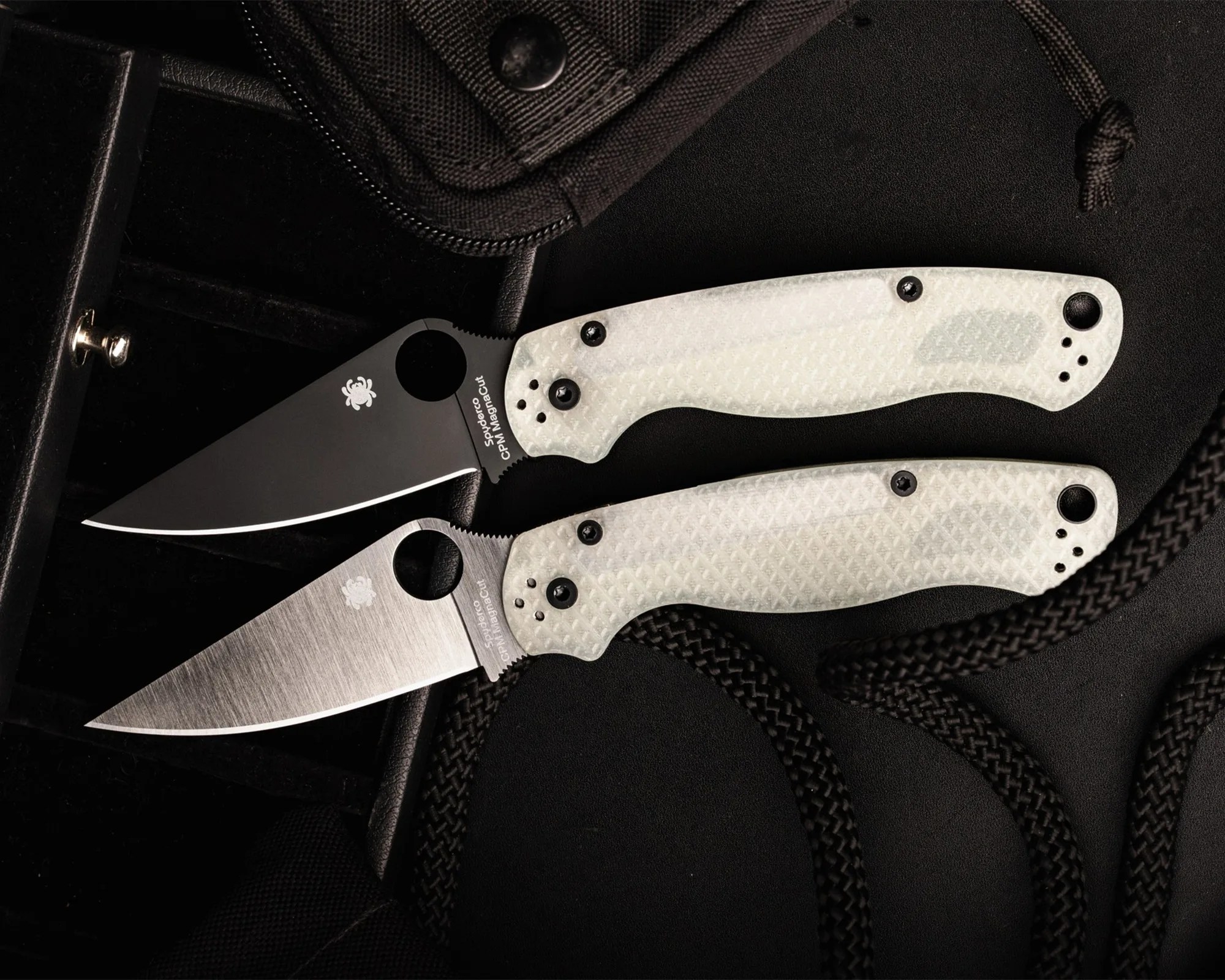 Two folding knives with white textured handles and one black-coated blade and one silver blade, placed on a black surface with black rope.