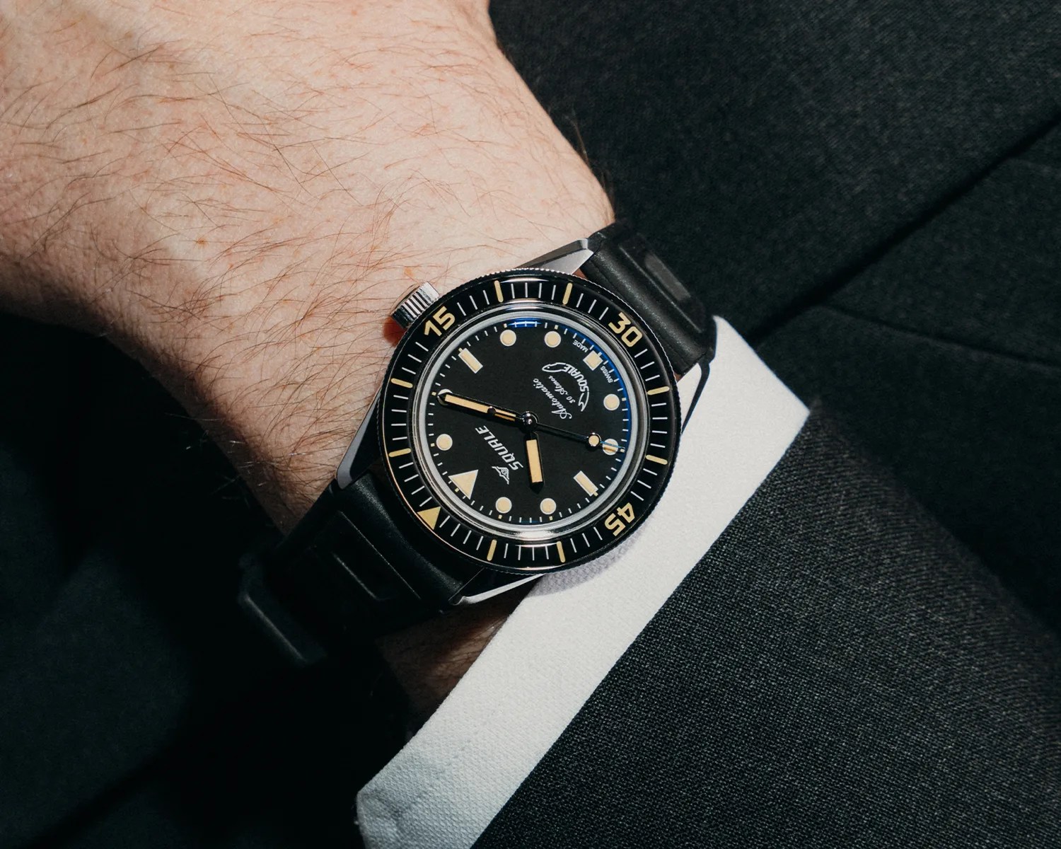 Black Squale dive watch with beige markers and black rubber strap worn on a wrist with a white shirt cuff and black suit.