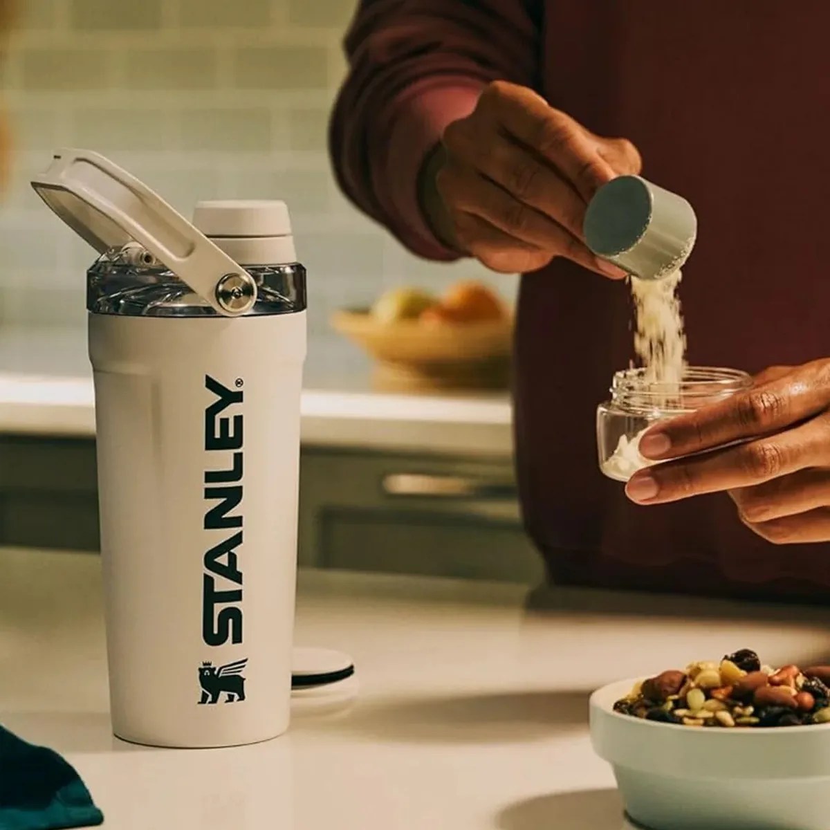 White Stanley insulated bottle with a flip-top lid on a kitchen counter next to a person scooping powder into a small container.