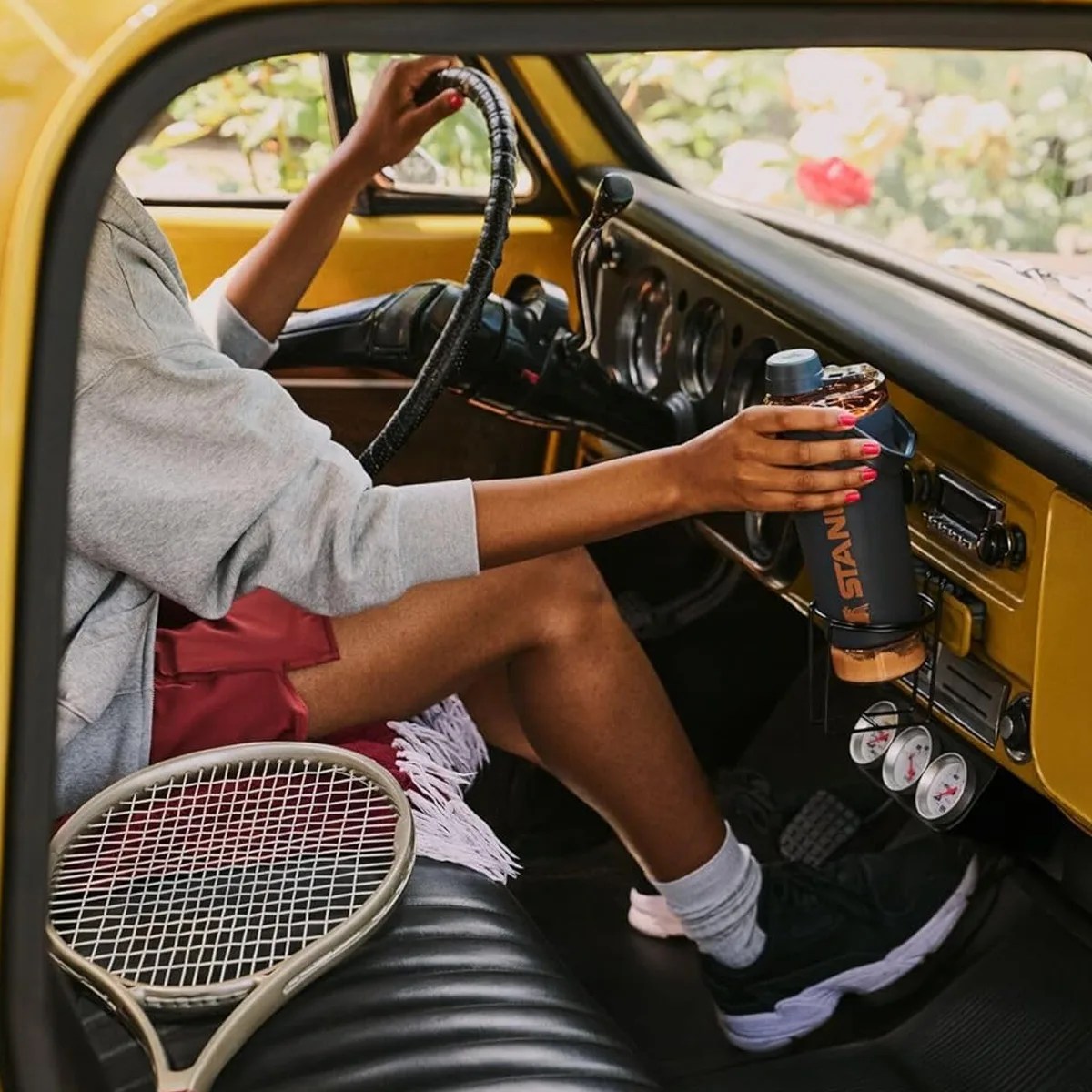Person in red shorts and gray sweatshirt holding a black Stanley water bottle inside a yellow car with a tennis racket on the seat.