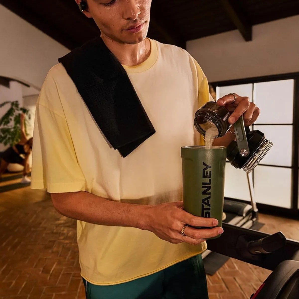 Person in a yellow shirt pouring powder into an olive green Stanley tumbler with a black towel over their shoulder.