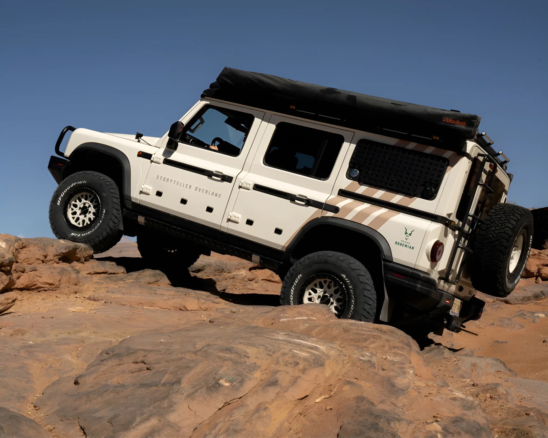 White off-road SUV with roof tent and spare tire climbing rocky terrain under clear blue sky.