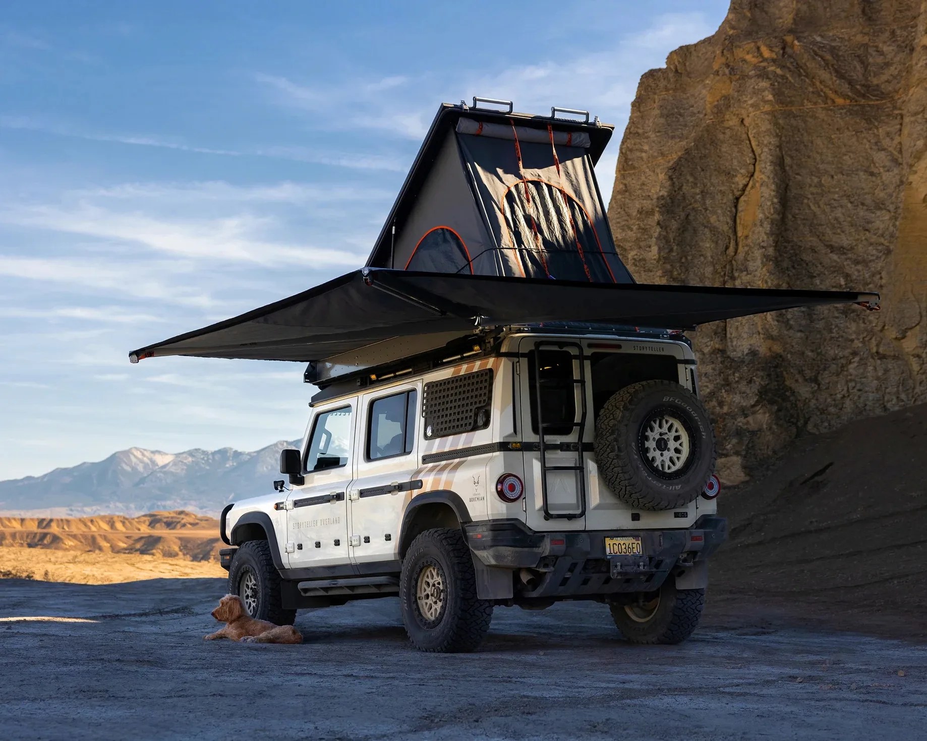 White off-road vehicle with rooftop tent and extended awning parked on rocky terrain with a dog lying nearby.