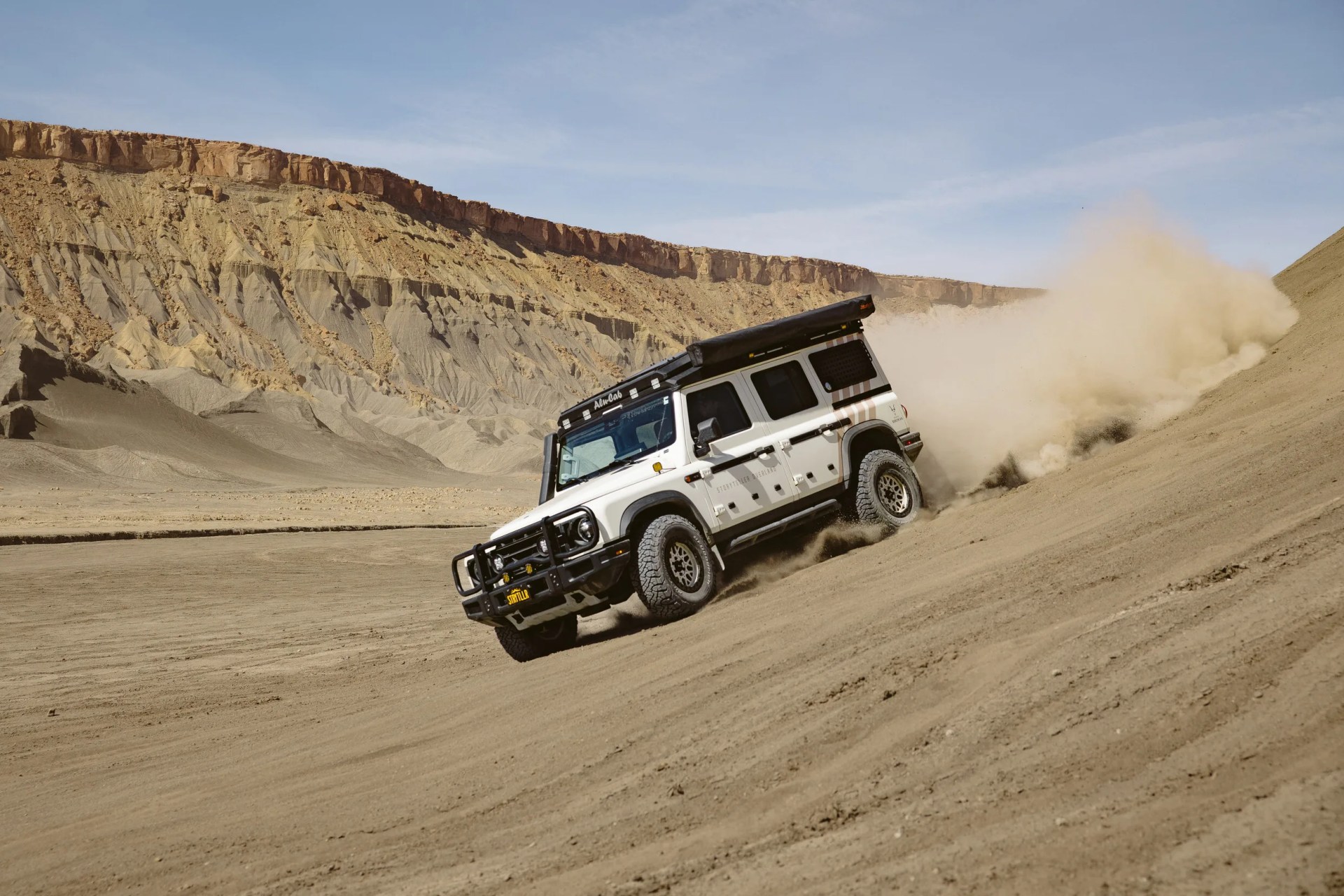 White off-road SUV with roof rack driving up a steep sandy hill in a desert landscape.