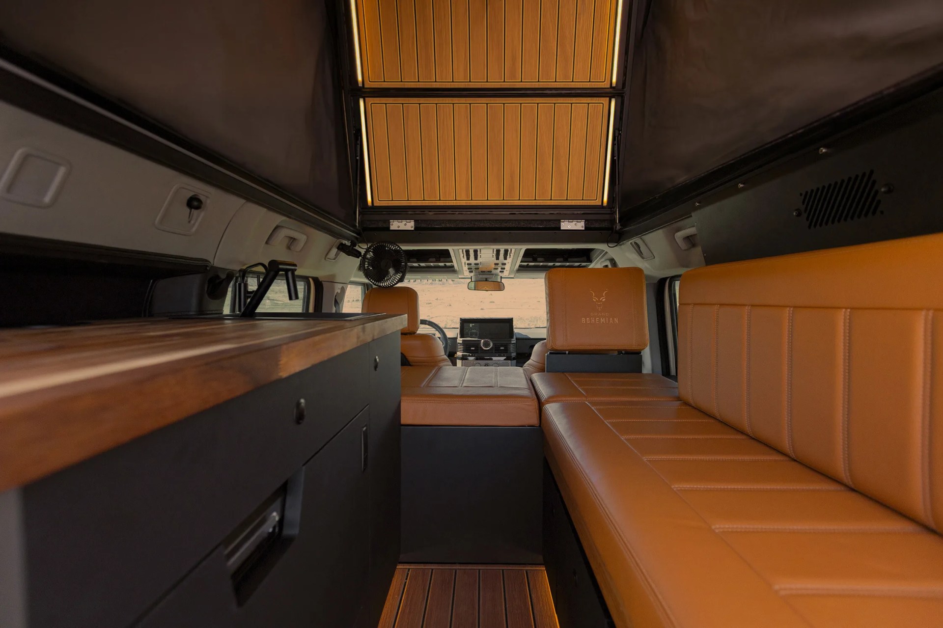 Interior of a camper van with tan leather seating, wooden countertop, and a partially open wooden roof panel.