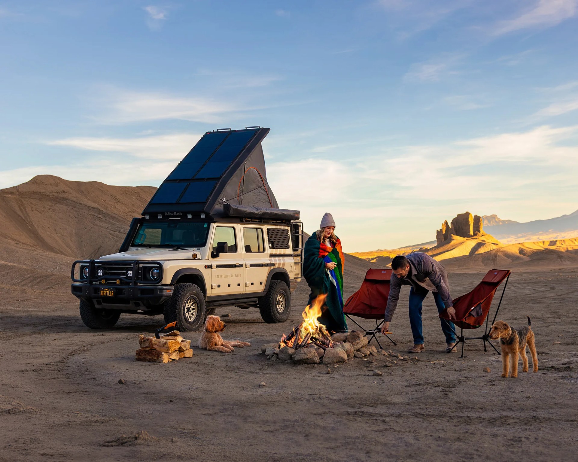 Beige off-road vehicle with rooftop tent and solar panels parked near a campfire with two people, two dogs, and camping chairs in a desert.