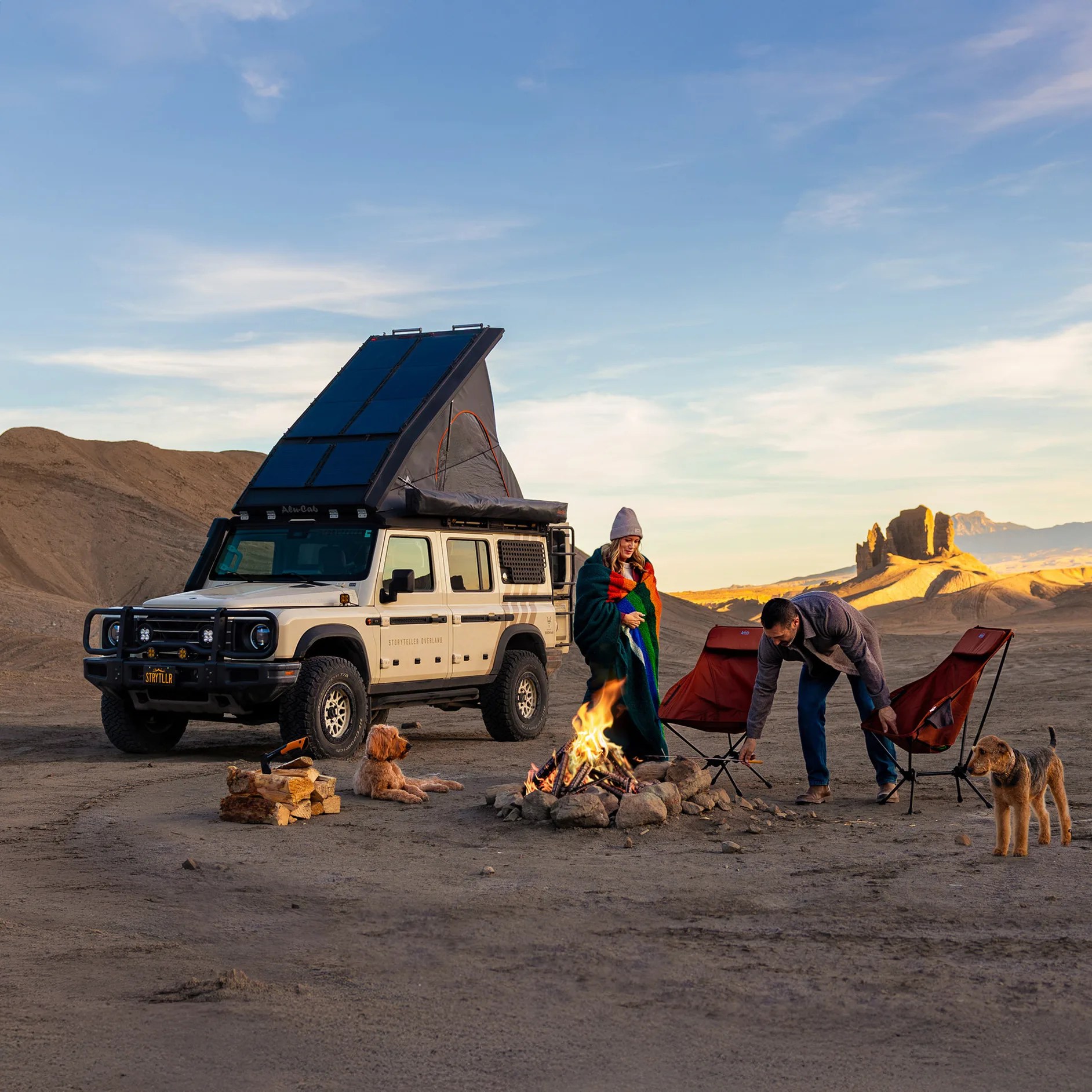 Beige off-road vehicle with rooftop tent and solar panels parked near a campfire with two people, two dogs, and camping chairs in a desert.