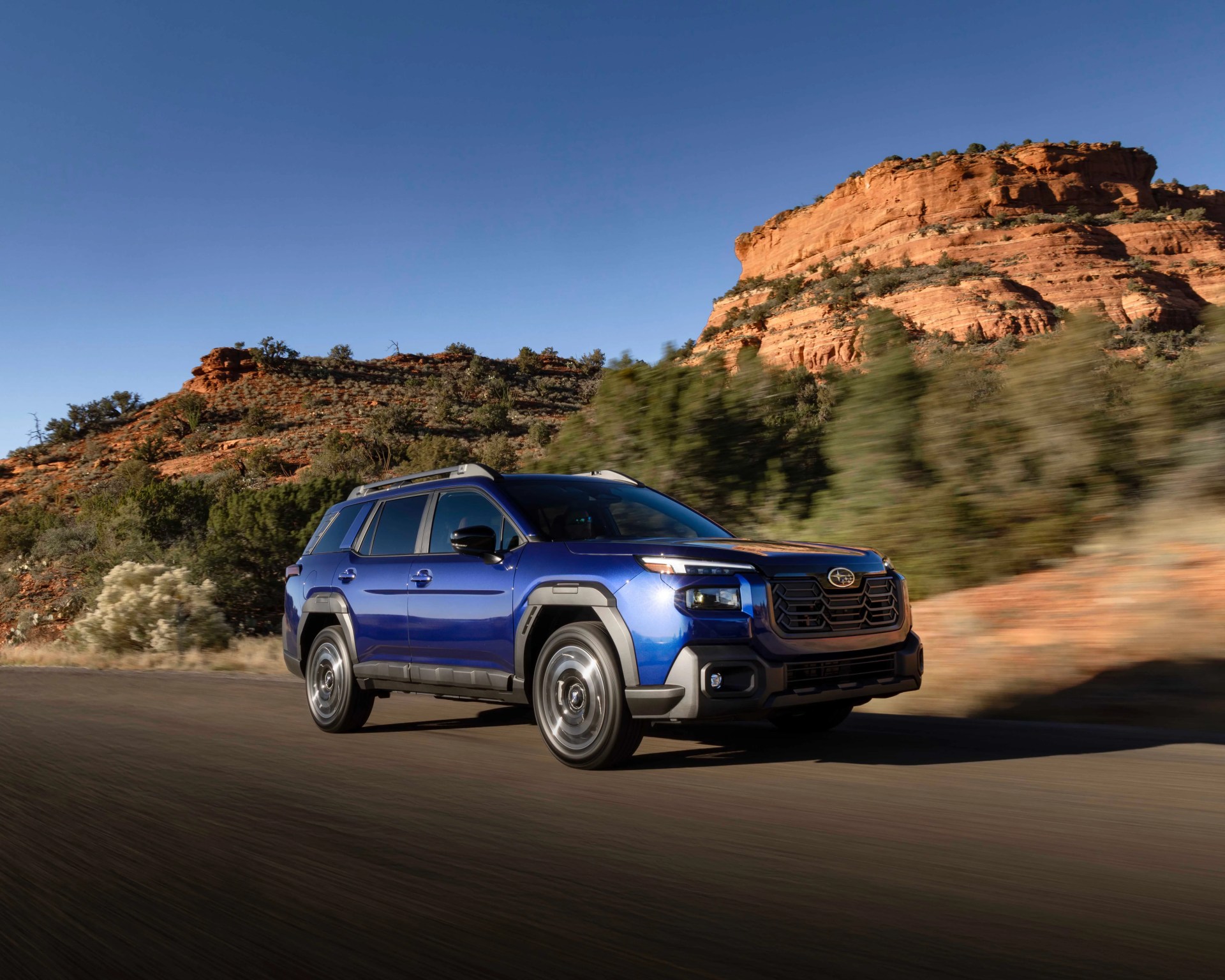 Blue Subaru SUV driving on a desert road with red rock formations and clear blue sky in the background.