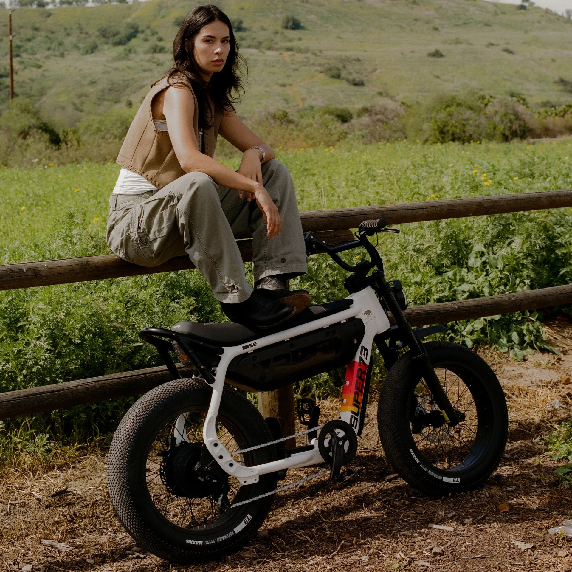 Person wearing a tan vest and cargo pants sitting on a white electric bike with "SUPER73" branding near a wooden fence in a green field.