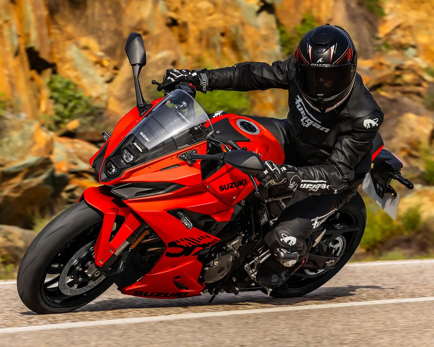 Rider in black gear leaning into a turn on a red Suzuki sport motorcycle on a paved road.