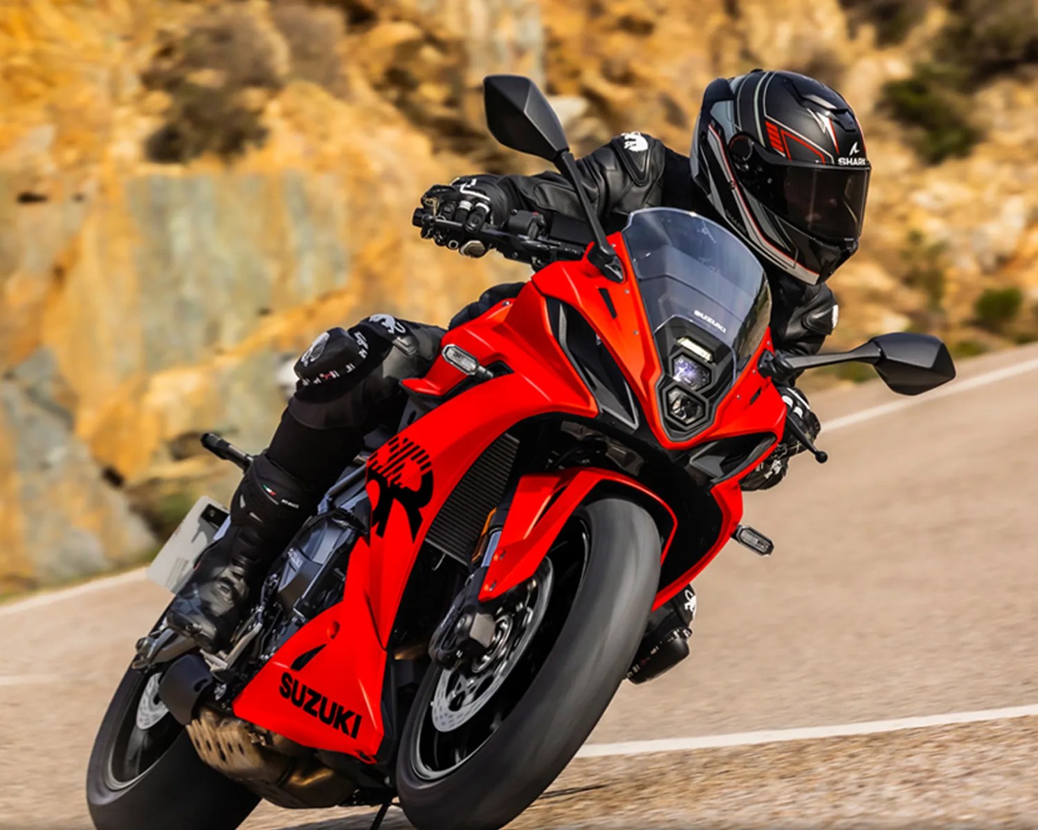 Red Suzuki sport motorcycle leaning into a curve on a mountain road with a rider in black gear and helmet.
