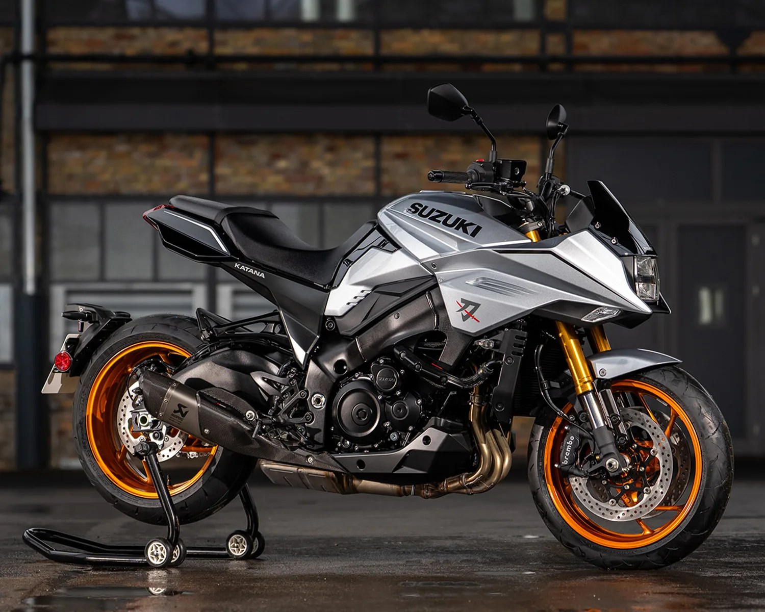 Silver Suzuki Katana motorcycle with orange wheels on a rear stand in an industrial setting.
