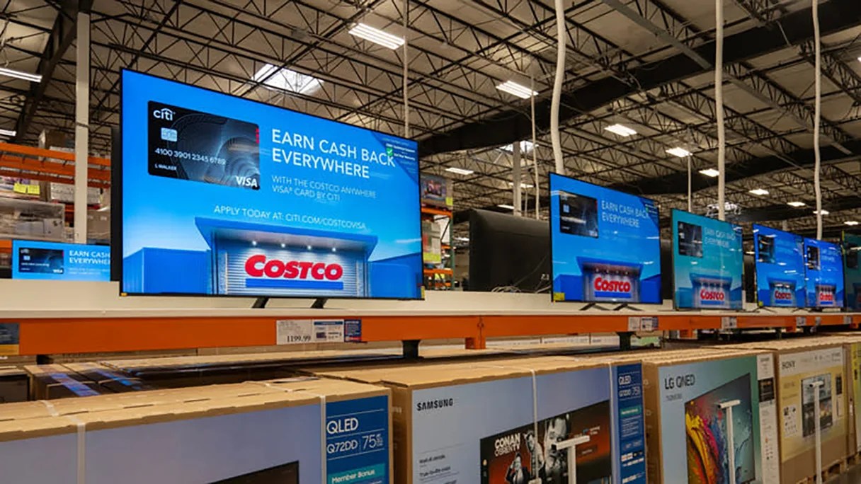 Row of large flat-screen TVs on display in a warehouse store with Costco credit card ads on screens.