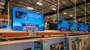 Row of large flat-screen TVs on display in a warehouse store with Costco credit card ads on screens.