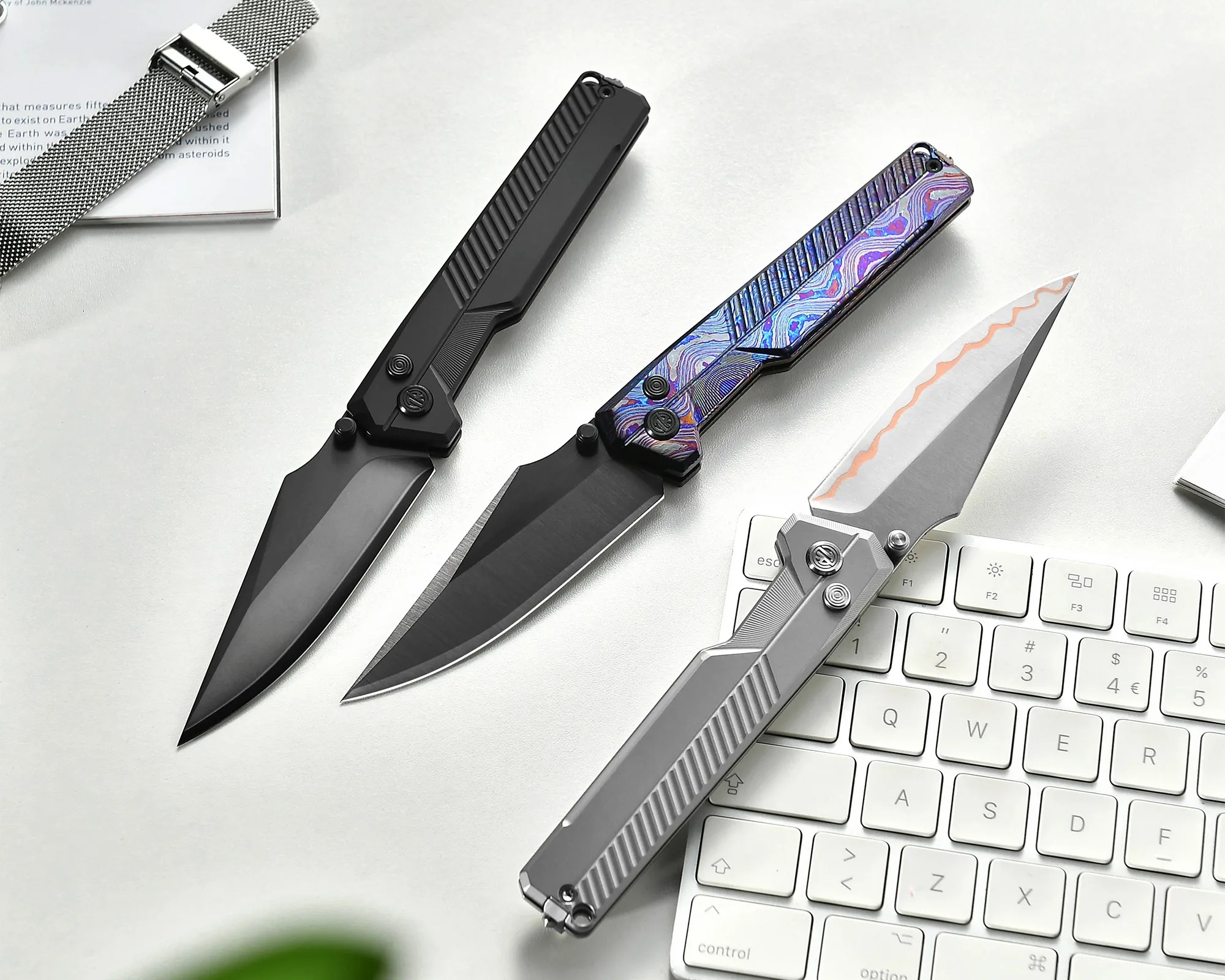Three folding knives with different handle finishes—black, iridescent purple-blue, and silver—laid on a white surface near a keyboard and a watch strap.