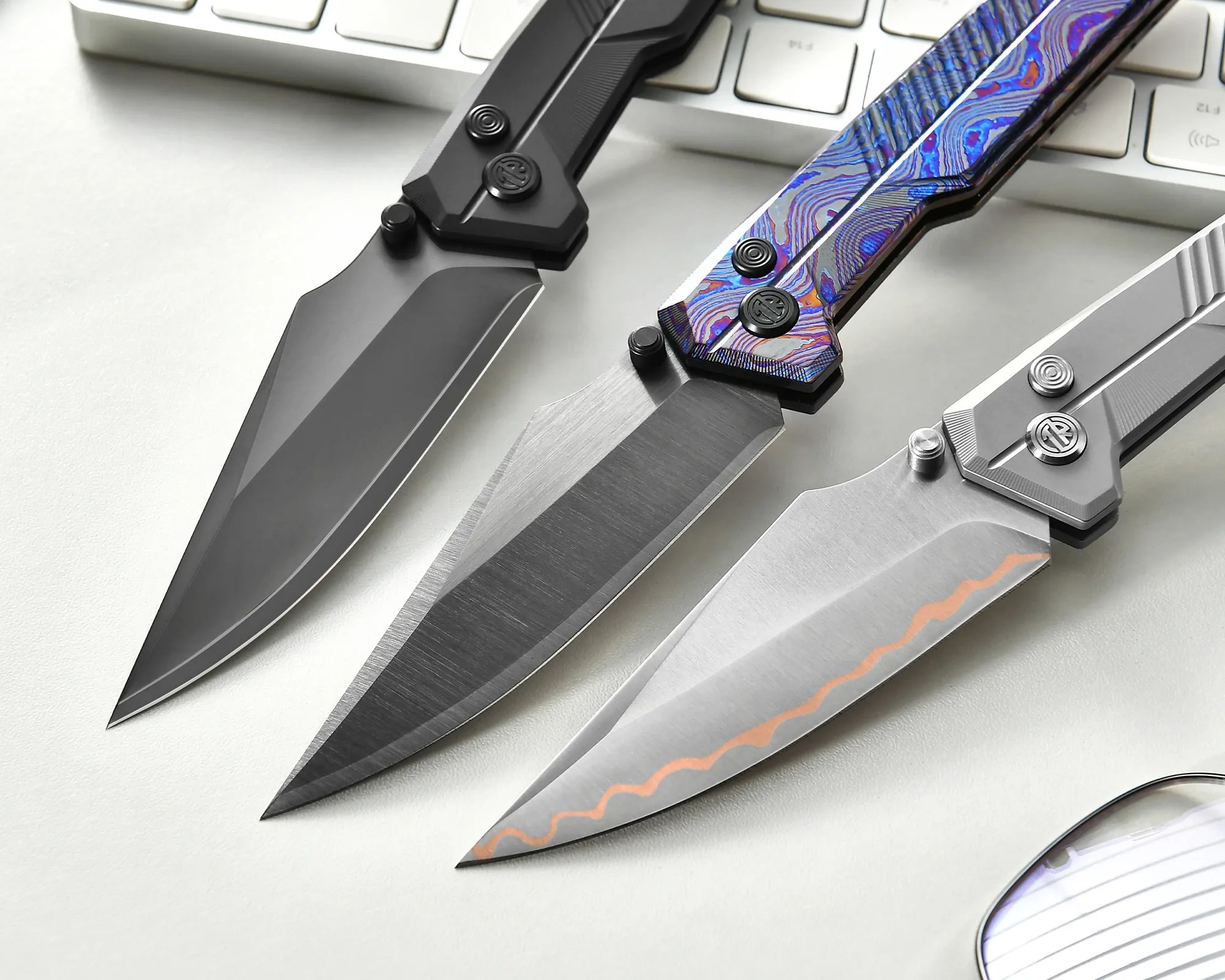 Three folding knives with distinct blade finishes—black, brushed steel, and silver with a wavy pattern—laid on a white surface near a keyboard.