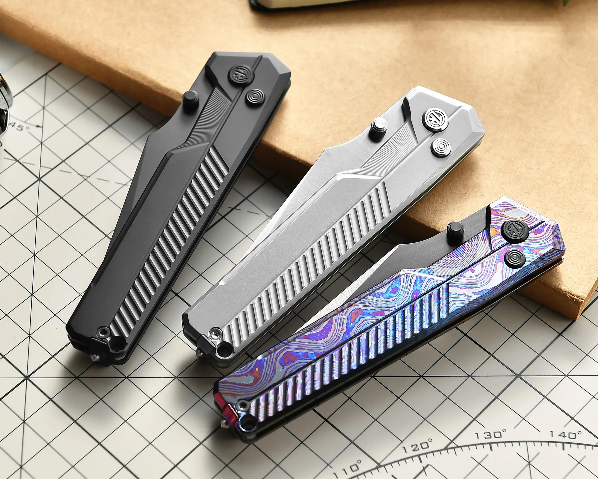Three closed folding knives with textured handles in black, silver, and iridescent blue-purple patterns on a grid cutting mat.