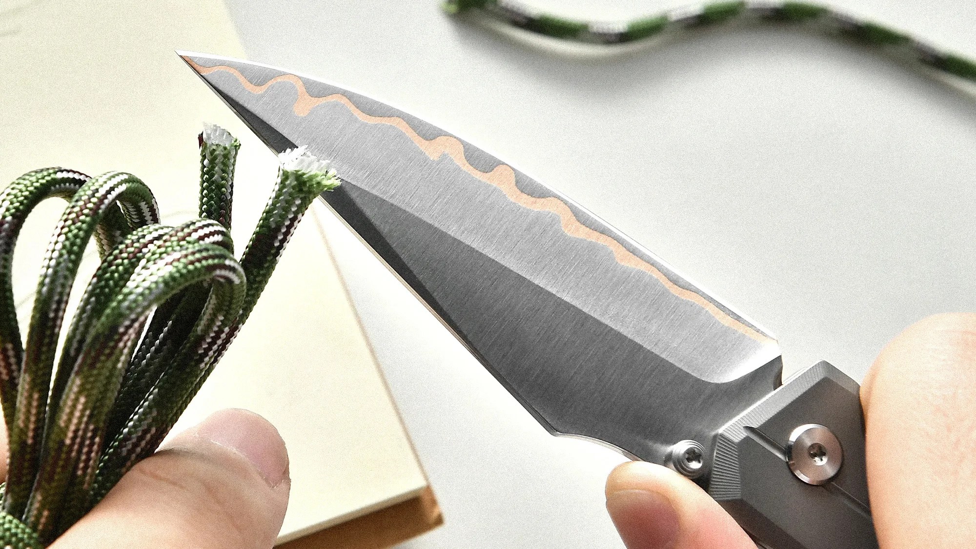 Close-up of a folding knife with a patterned blade cutting green and white paracord held in two hands.