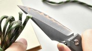 Close-up of a folding knife with a patterned blade cutting green and white paracord held in two hands.