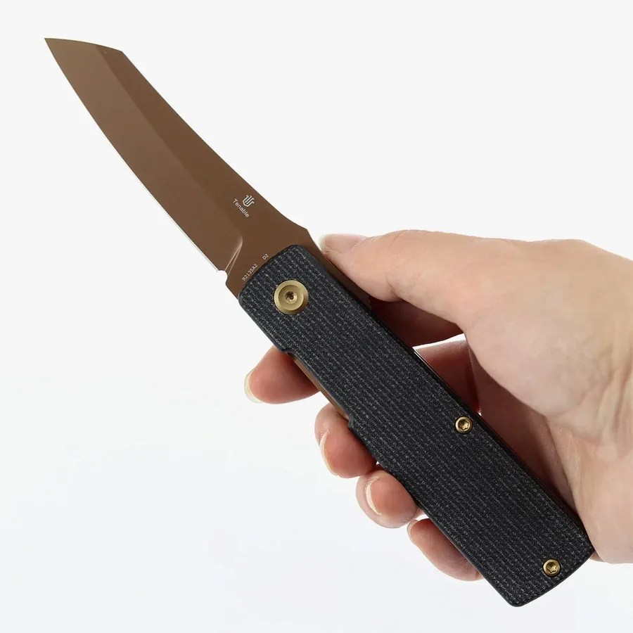 Hand holding a folding knife with a brown blade and textured black handle with brass screws.
