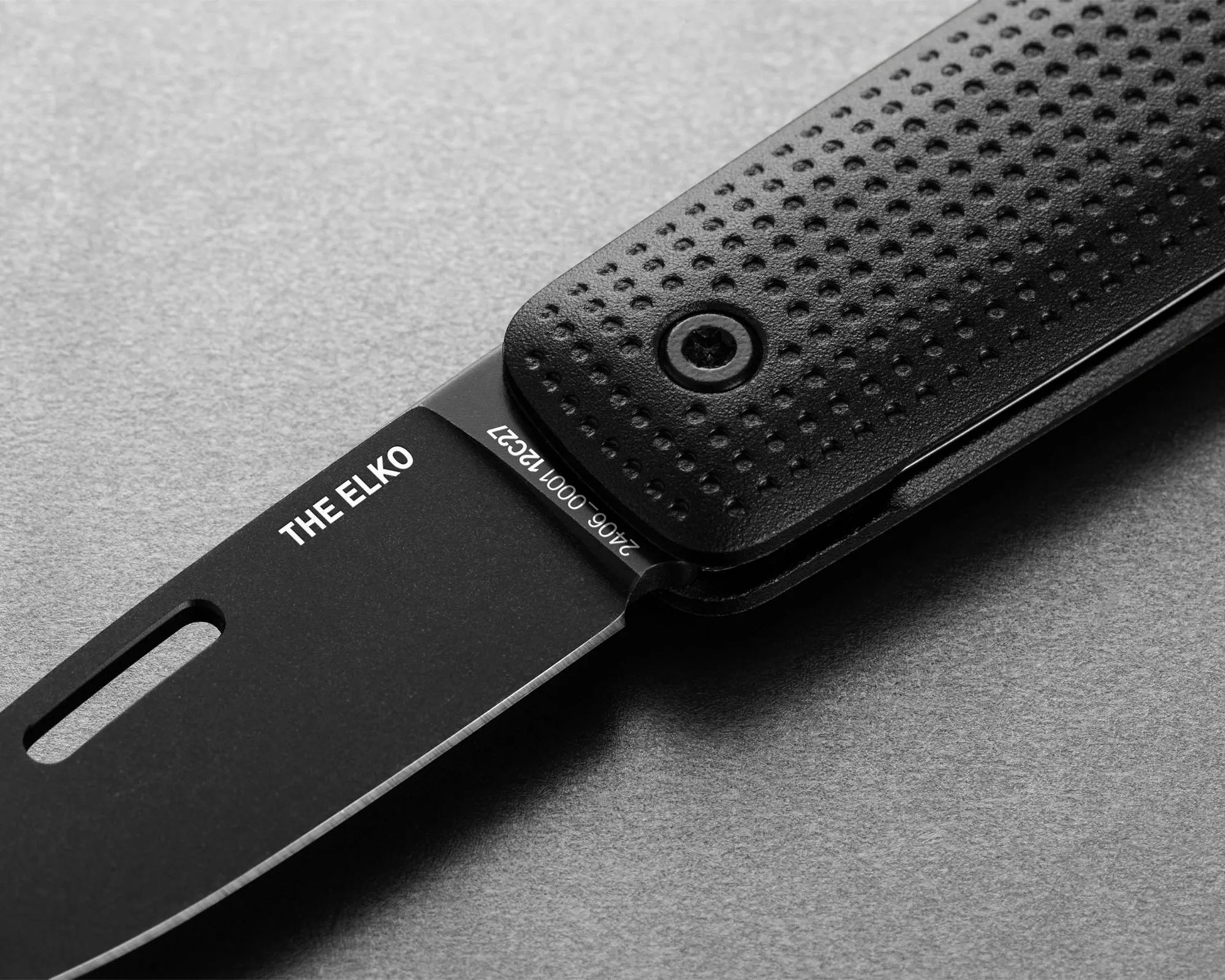 Black folding knife blade labeled "THE ELKO" with textured black handle on gray surface.