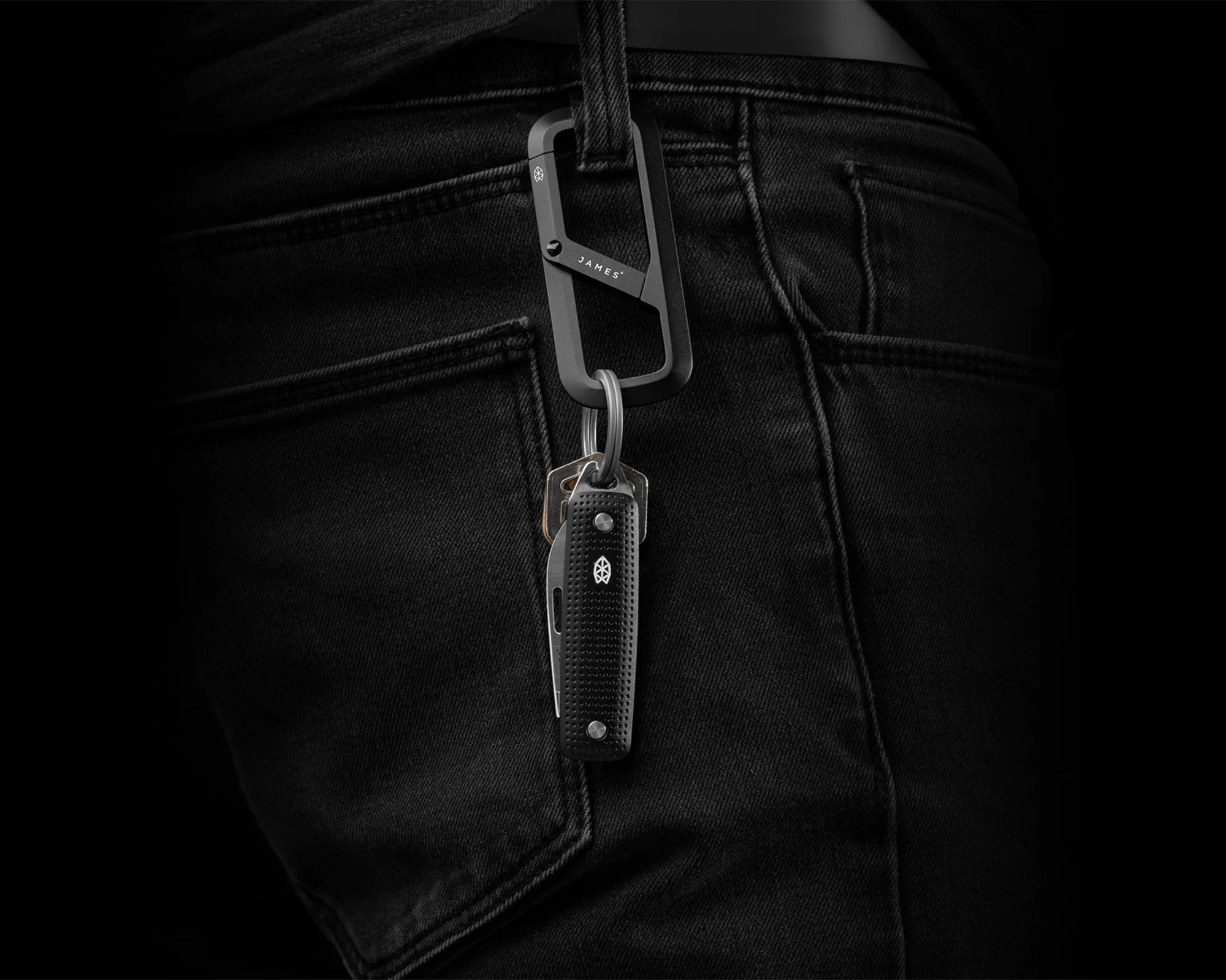 Black textured pocket knife and keys attached to a black metal carabiner clipped to a black denim pocket.