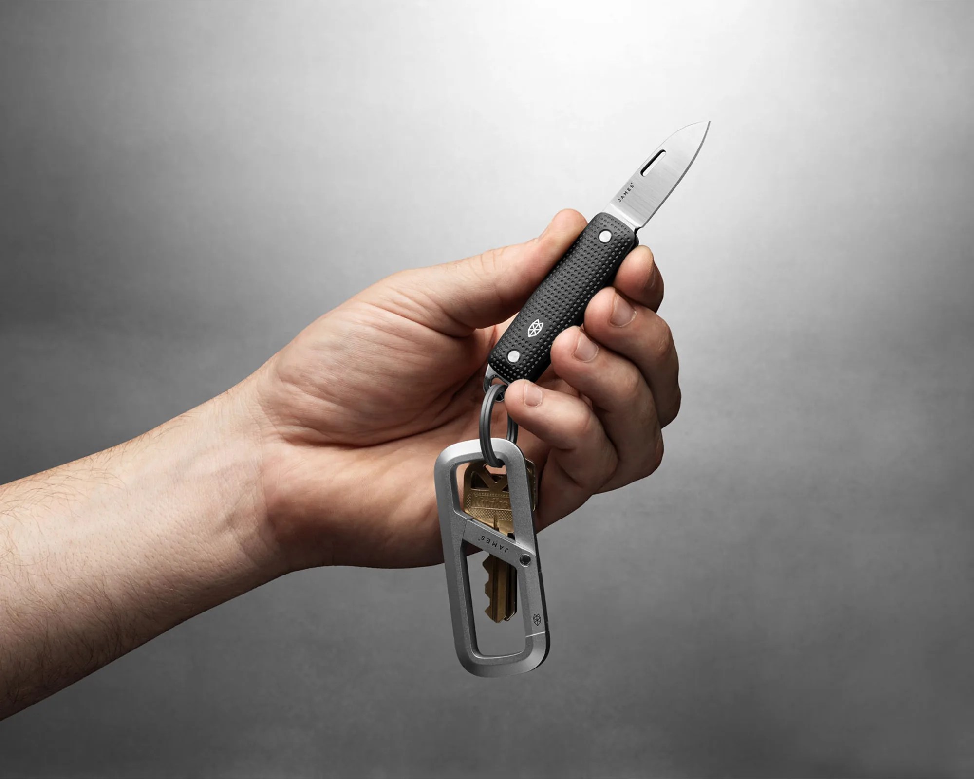 Hand holding a small black folding knife attached to a keyring with a silver carabiner and keys.