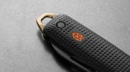 Black textured folding knife handle with an orange geometric logo and a brass loop on a gray surface.