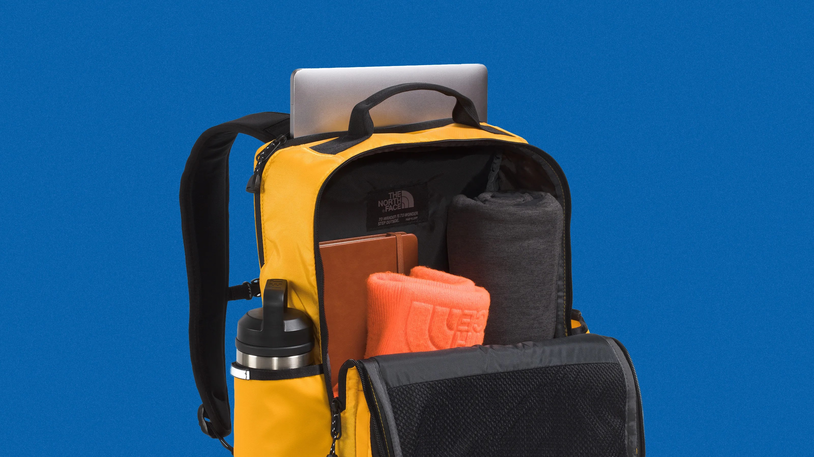 Yellow North Face backpack with black straps, containing a laptop, orange and gray rolled clothes, a brown notebook, and a water bottle in a side pocket.