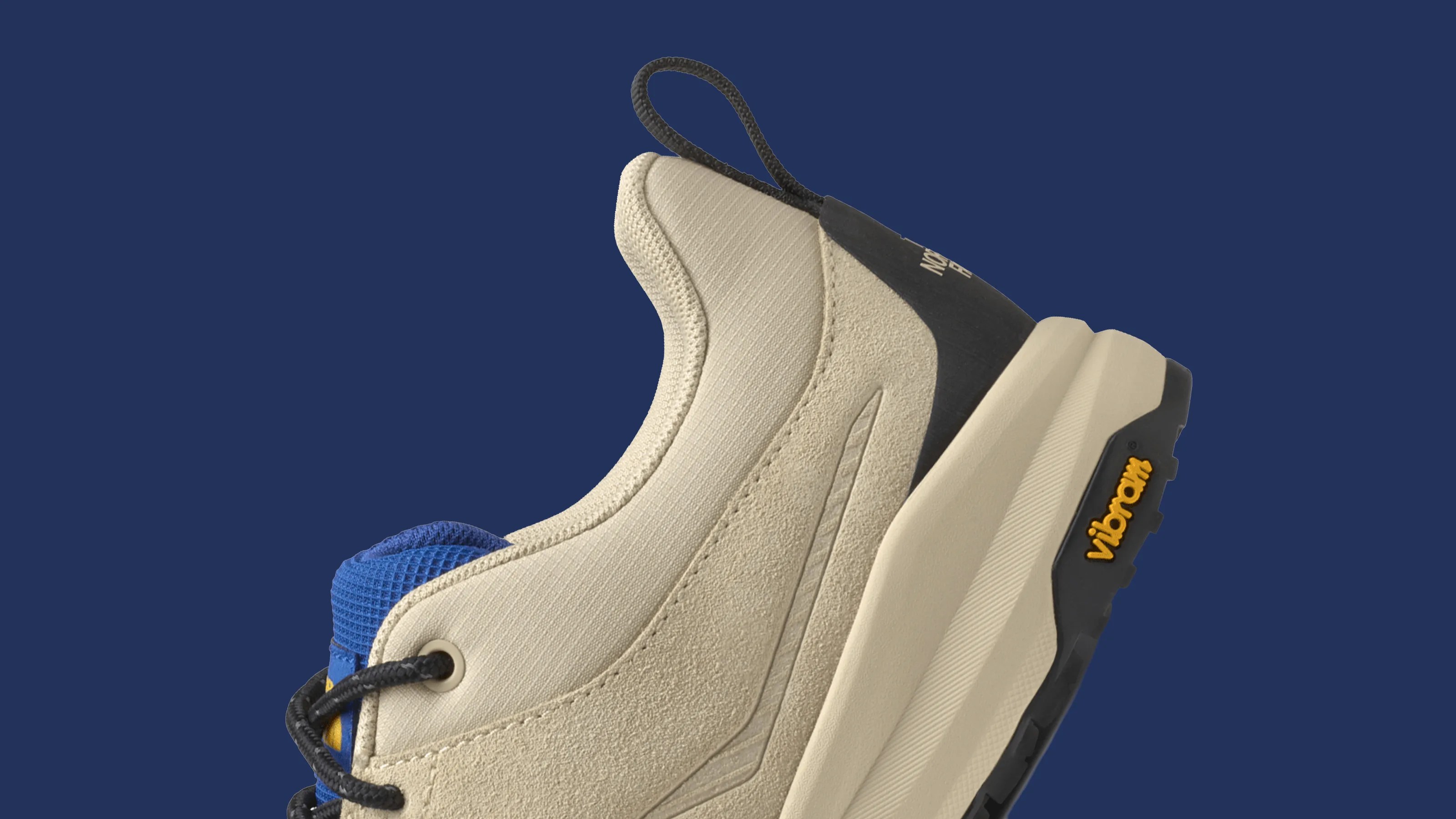 Beige and blue sneaker with black laces and Vibram sole on a dark blue background.