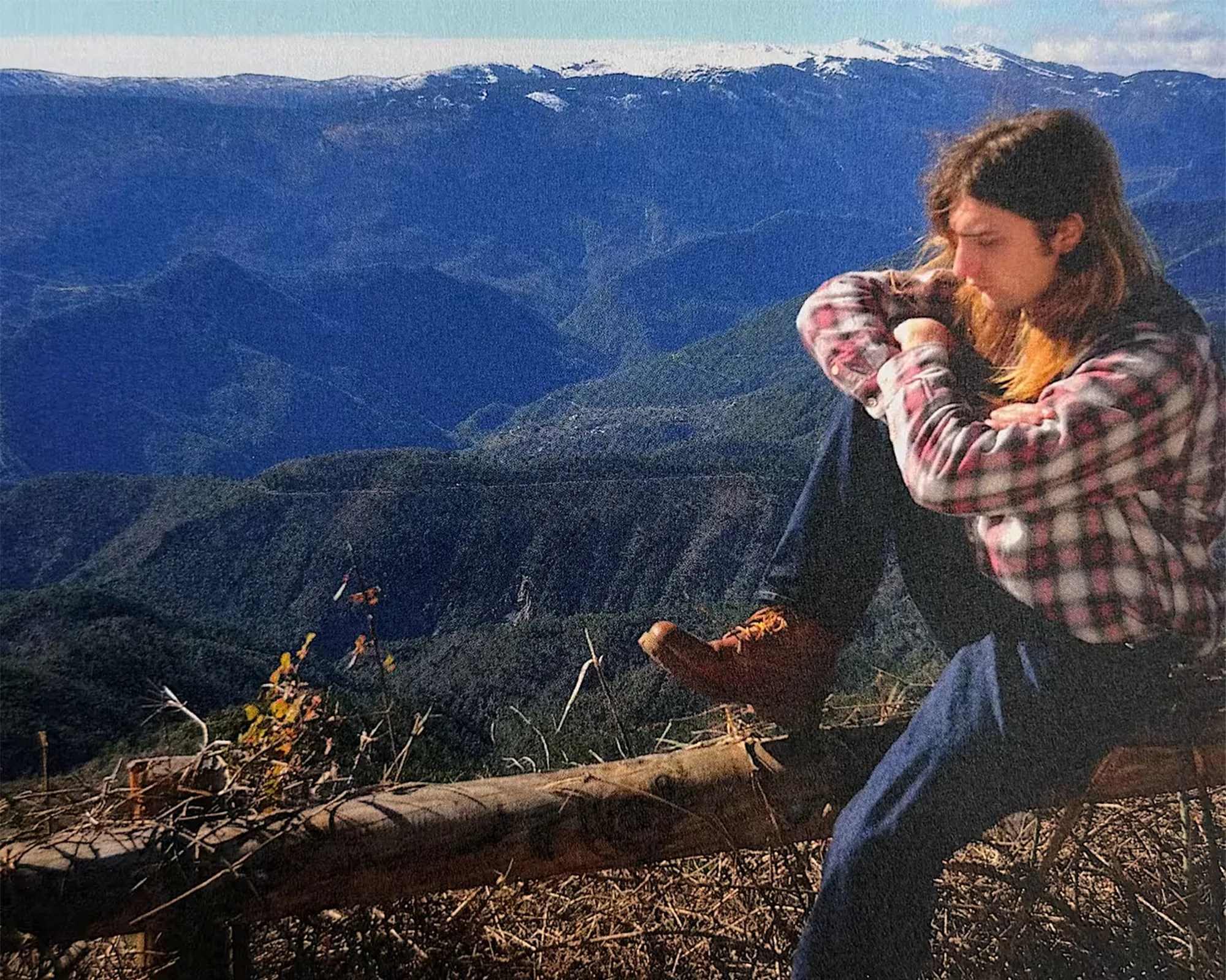 Person in a red and white plaid shirt and brown boots sitting on a log with snow-capped mountains in the background.