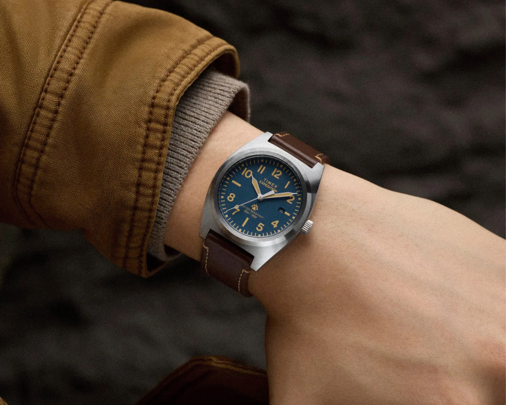 Wristwatch with blue dial, yellow numerals, silver case, and brown leather strap worn on a wrist with a brown jacket sleeve.