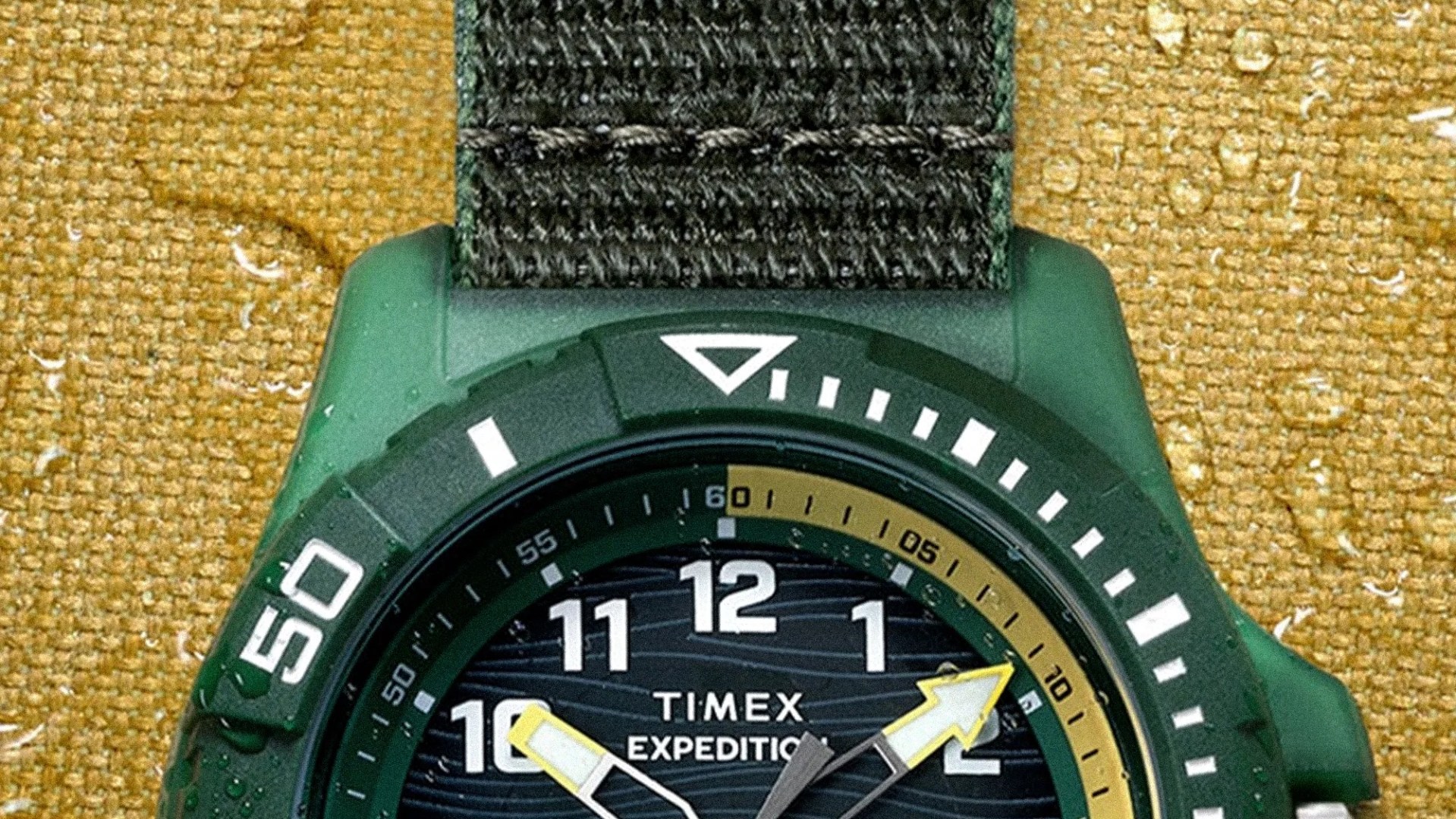 Green Timex Expedition watch with black fabric strap and yellow-accented hands on a textured yellow surface with water droplets.