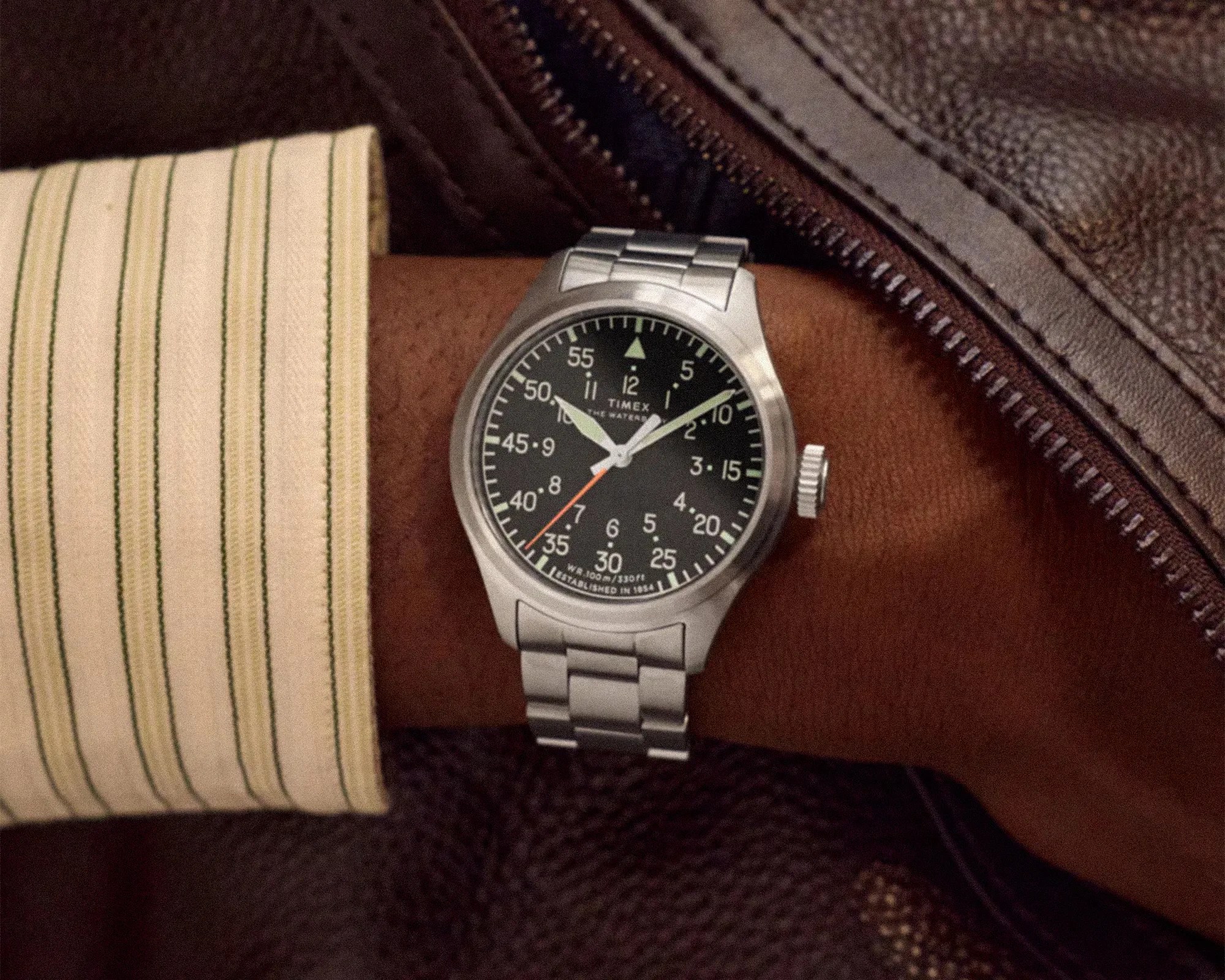 Silver Timex wristwatch with black dial and white numerals on a wrist, next to a brown leather jacket.